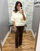 A woman takes a mirror selfie in a fitting room wearing a cream short-sleeve knit top and dark brown wide-leg pants with tan shoes.