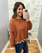 A woman takes a mirror selfie wearing a rust-orange knit sweater with white ribbed trim at the crew neck and scalloped hem, paired with dark blue jeans.