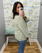 A woman takes a mirror selfie wearing a long-sleeve, wide-leg striped shirt with blue jeans. The shirt features thin vertical stripes in olive green and white.