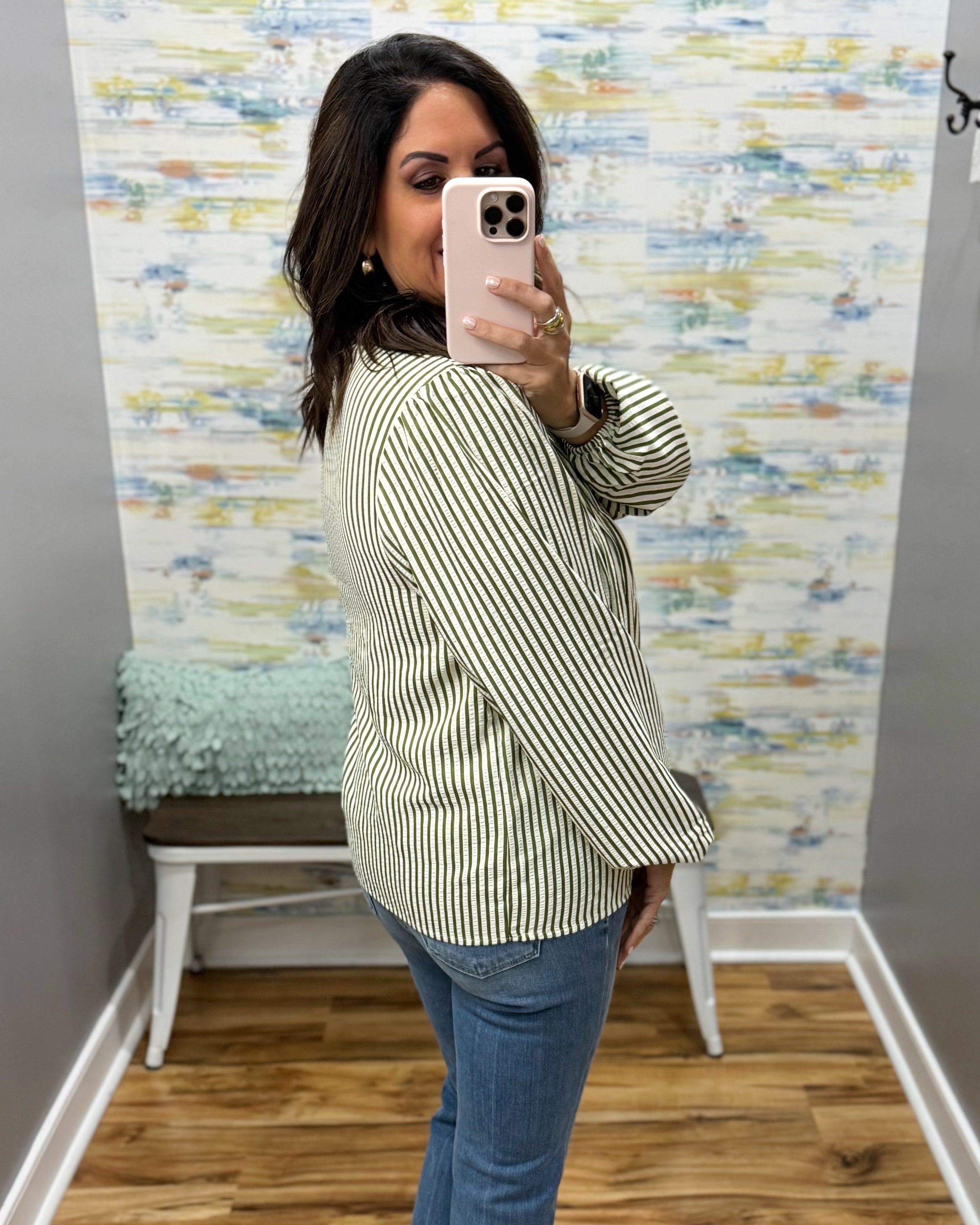 A woman takes a mirror selfie wearing a long-sleeve, wide-leg striped shirt with blue jeans. The shirt features thin vertical stripes in olive green and white.