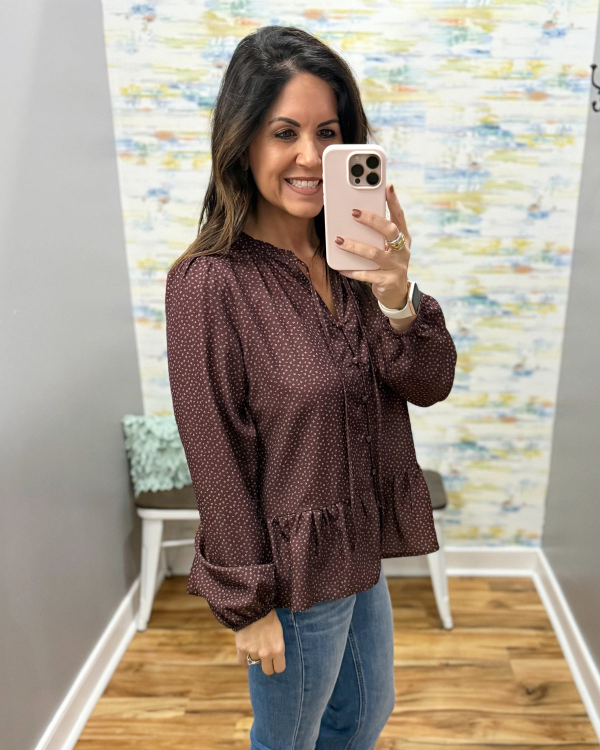 A woman takes a mirror selfie wearing a dark brown long-sleeve blouse with a small white geometric pattern, paired with blue jeans.