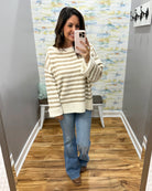 A woman takes a mirror selfie in a fitting room wearing a cream and tan striped knit sweater, blue flare jeans, and brown boots.