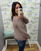 A woman takes a mirror selfie wearing a taupe brown knit sweater and blue jeans in a fitting room with patterned wallpaper and a bench.