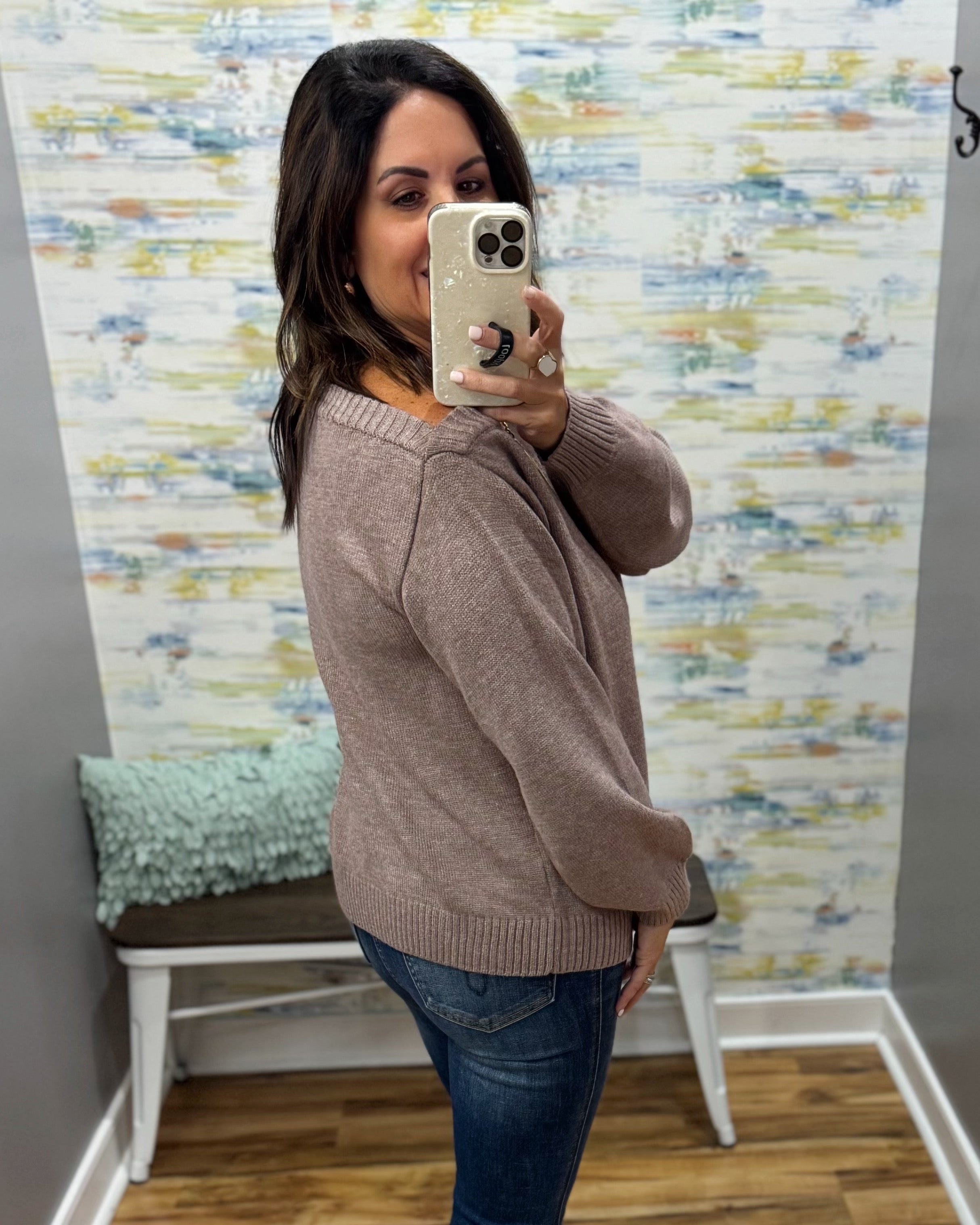 A woman takes a mirror selfie wearing a taupe brown knit sweater and blue jeans in a fitting room with patterned wallpaper and a bench.