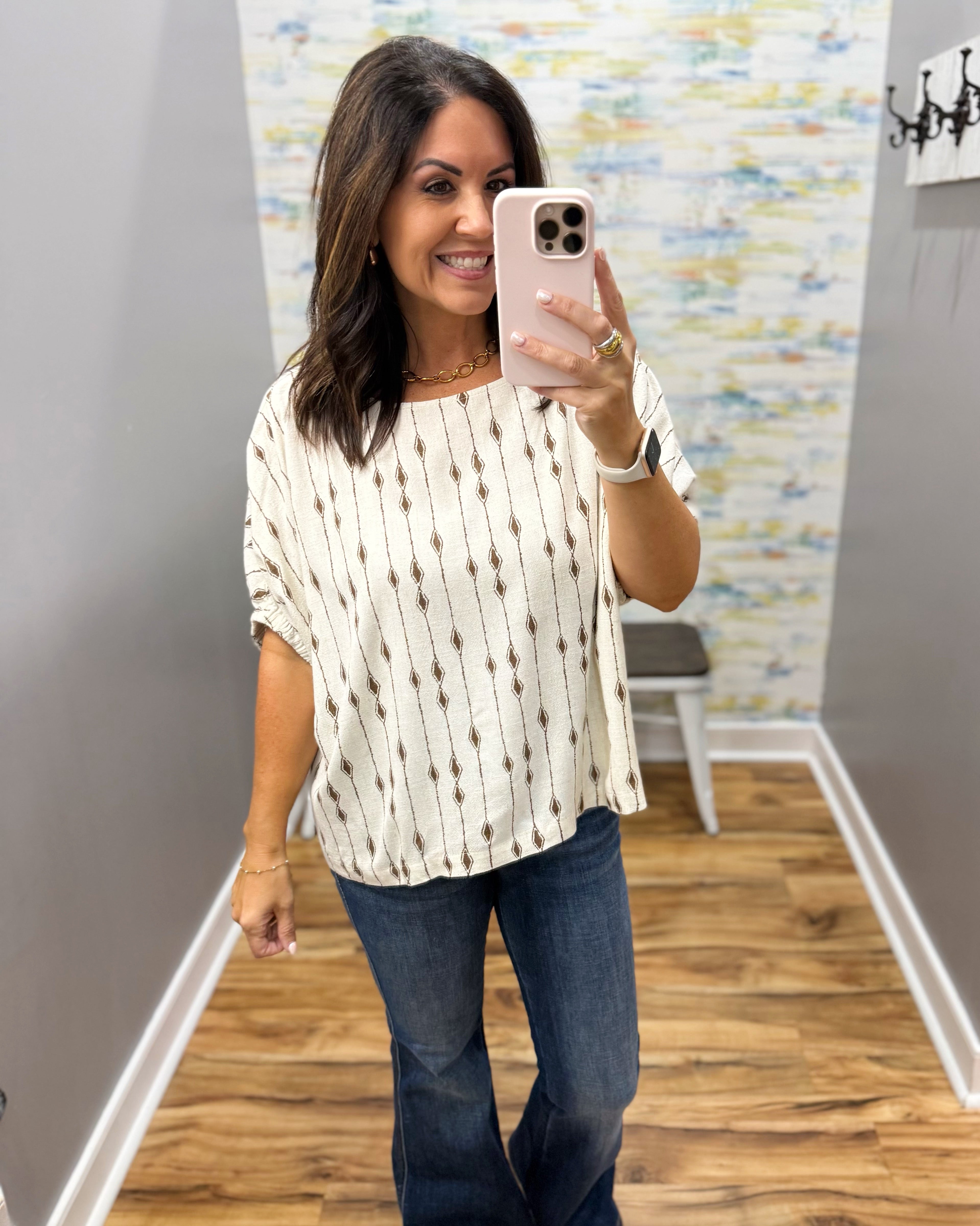 A woman takes a mirror selfie in a boutique showing a cream-colored short-sleeve top with a vertical brown geometric pattern, paired with dark blue jeans.