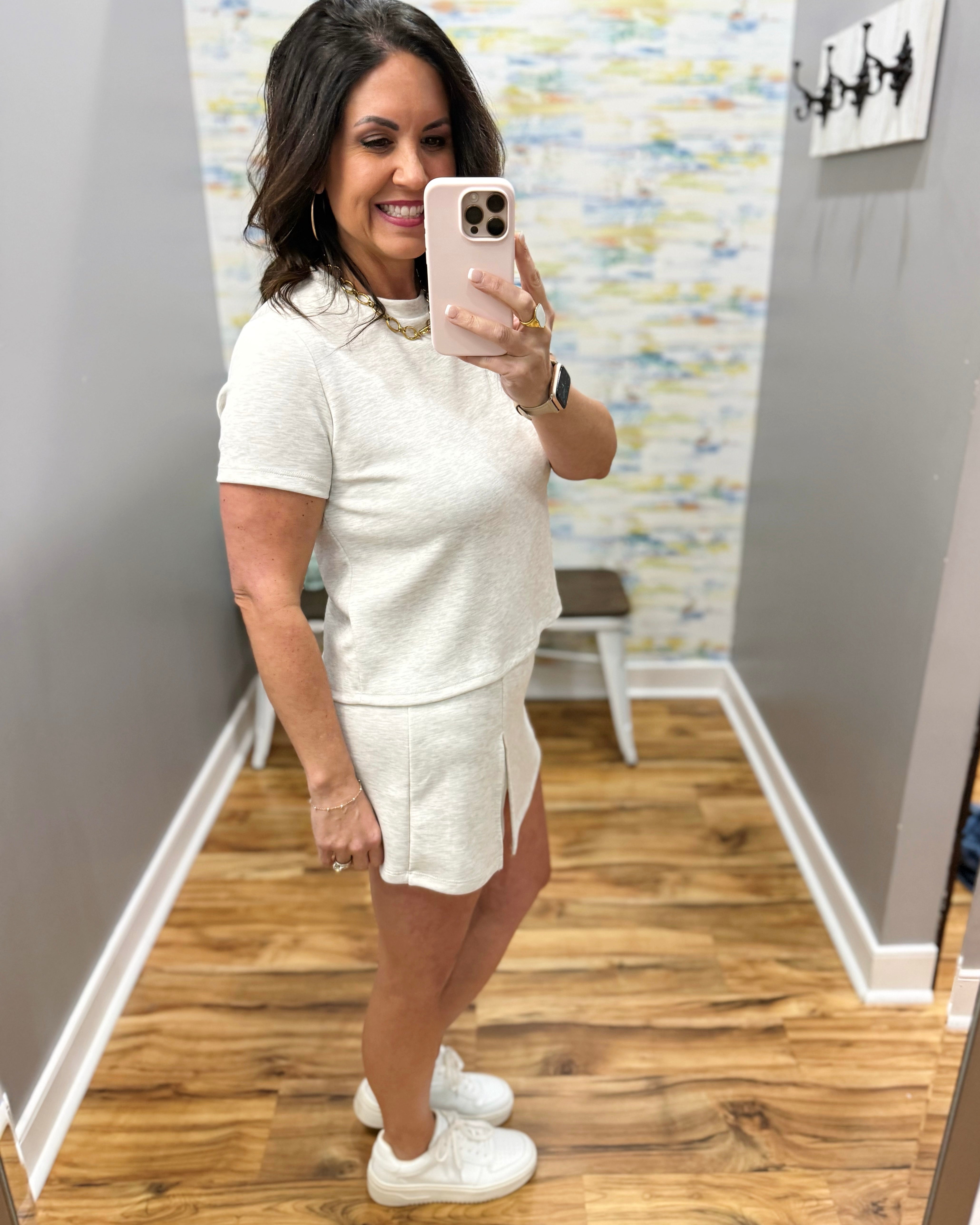 Woman taking a mirror selfie wearing a white outfit in a room with wooden flooring and a colorful wall.