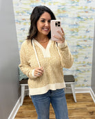 A woman takes a mirror selfie wearing a cream and tan marled knit sweater with a V-neck and drawstrings, paired with blue jeans in an indoor room setting.