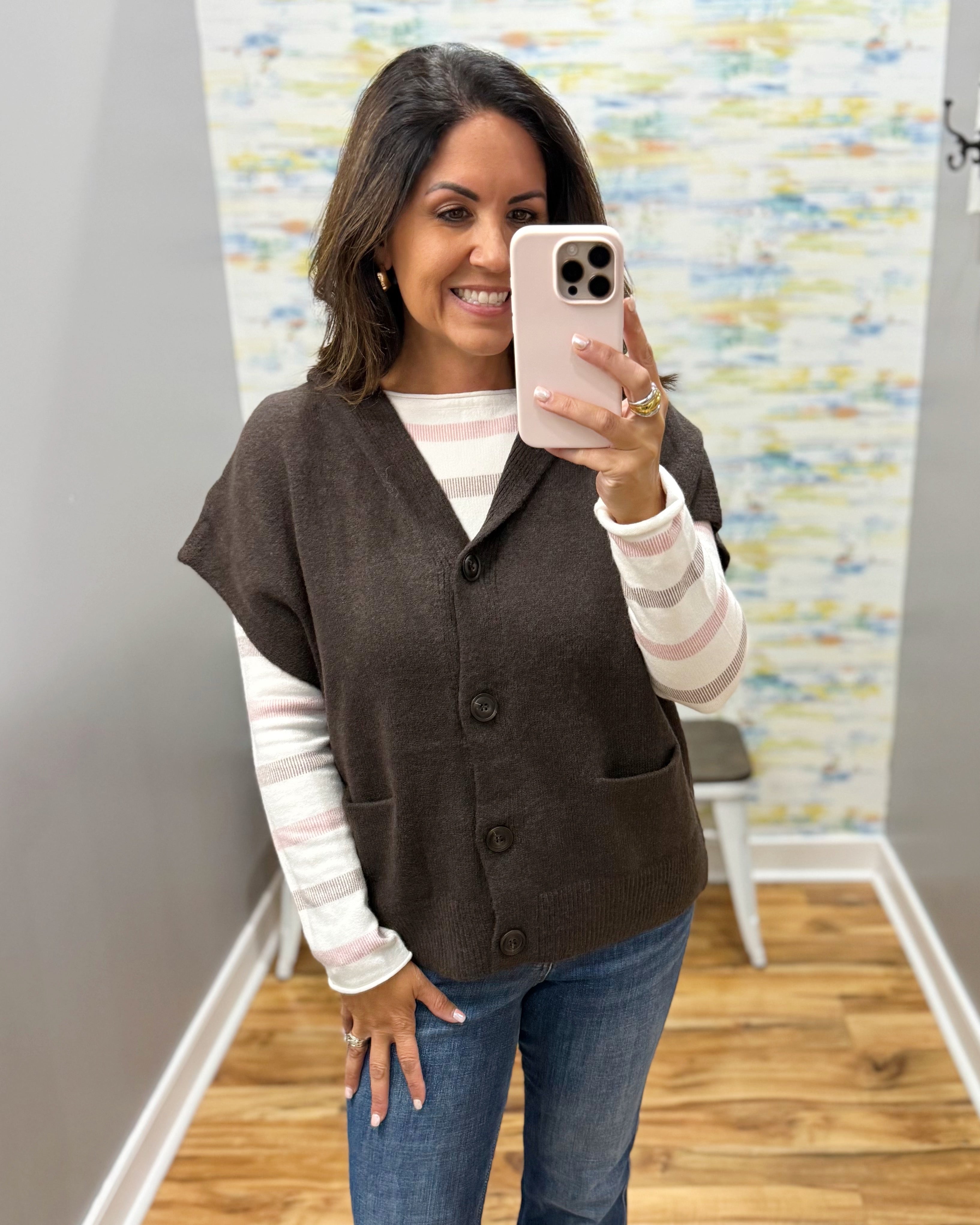 A woman taking a mirror selfie wearing a brown button-up knit vest over a pink and white striped long-sleeve shirt, paired with blue jeans.