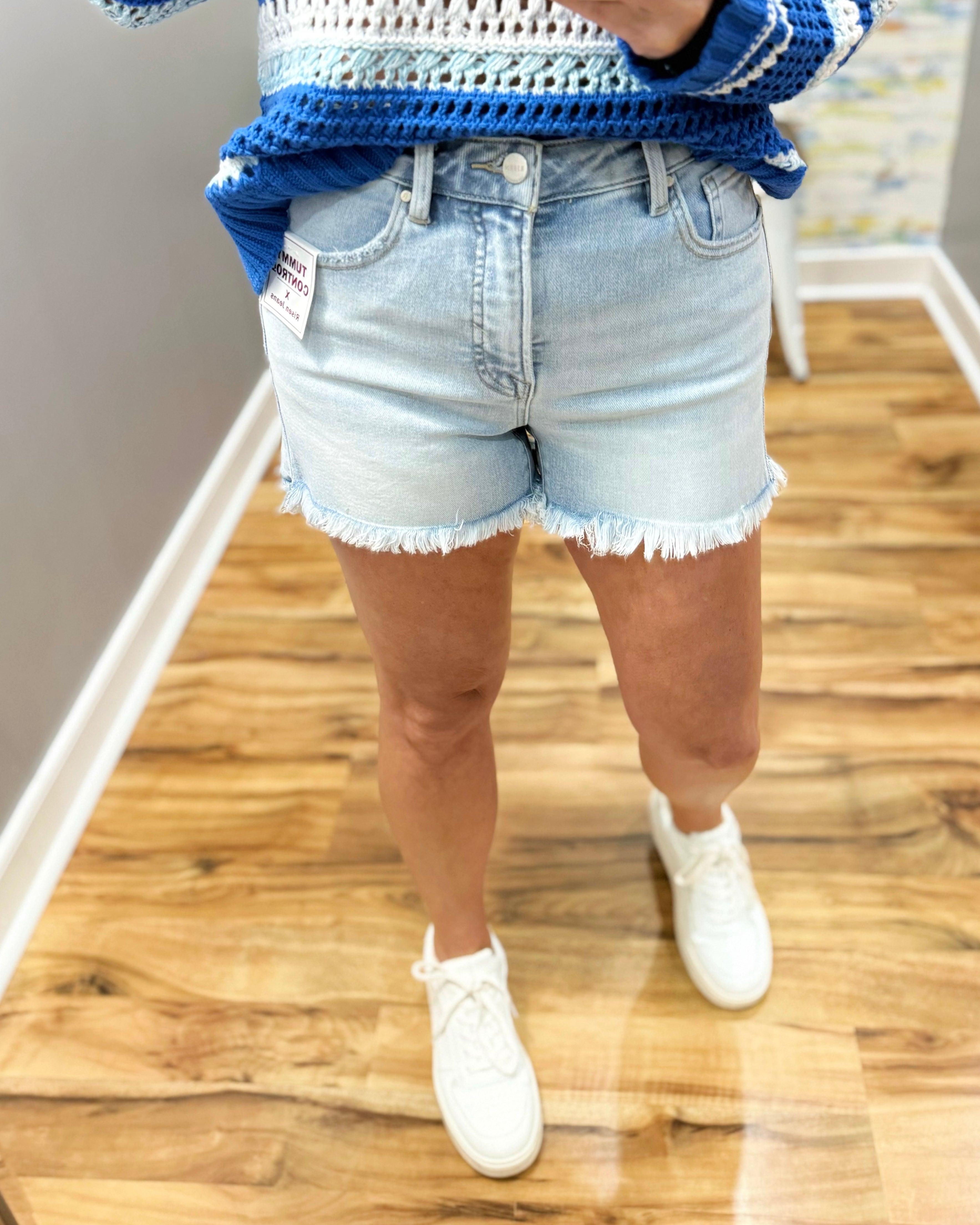 Person wearing a blue and white striped sweater, light denim shorts, and white sneakers on a wooden floor.