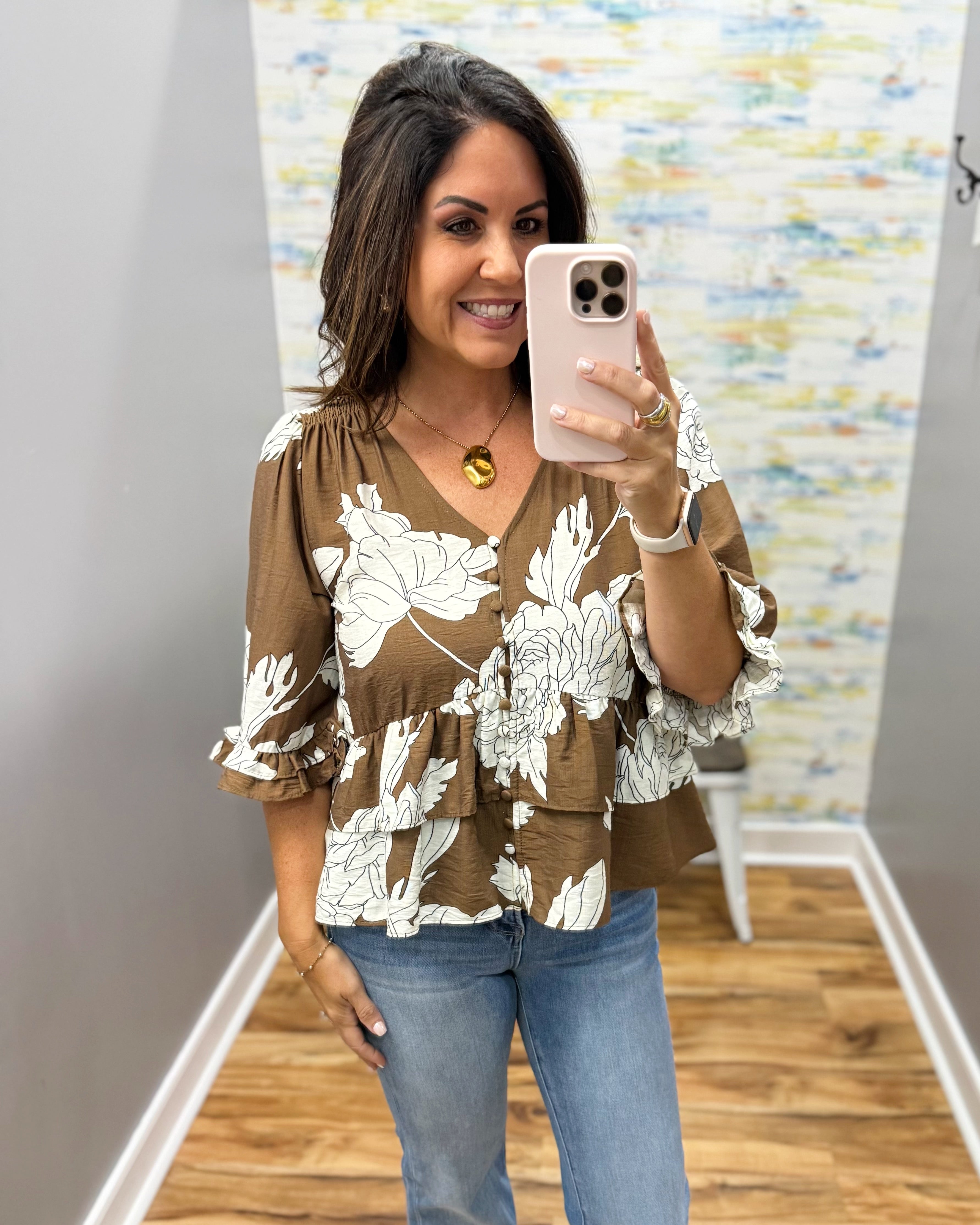 A woman takes a mirror selfie wearing a brown and white floral print blouse with tiered ruffle sleeves, paired with blue jeans, in a fitting room.