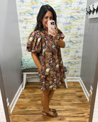 A woman takes a mirror selfie in a fitting room wearing a short puff-sleeve dress with an all-over floral print in earthy tones of brown, burgundy, green, and cream. She is wearing brown flat sandals and holding a light pink smartphone.