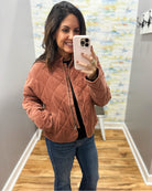 A woman takes a mirror selfie wearing a dusty rose quilted zip-up jacket, blue jeans, and a black top in a home hallway.