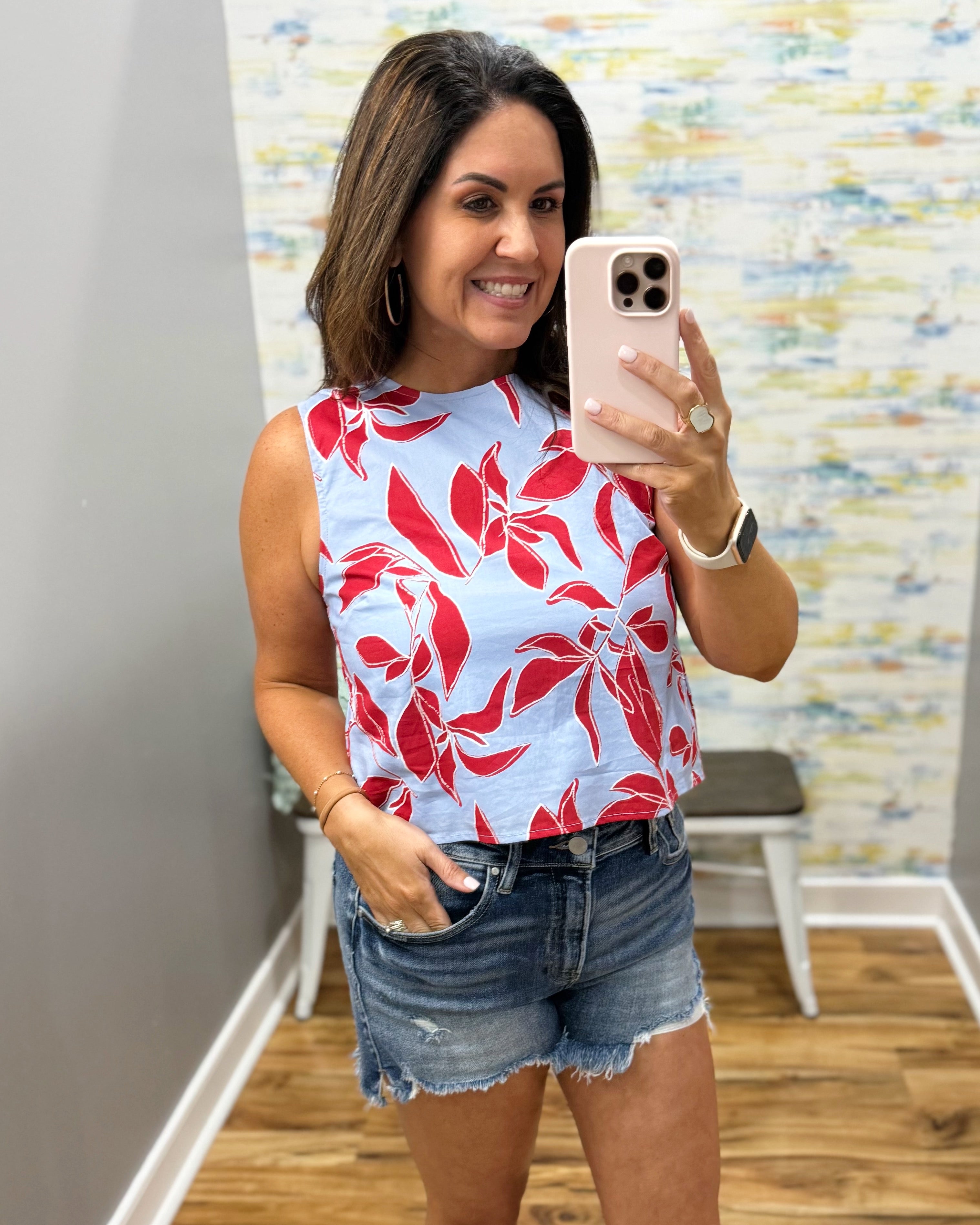 Woman taking a mirror selfie wearing a red floral top and denim shorts.