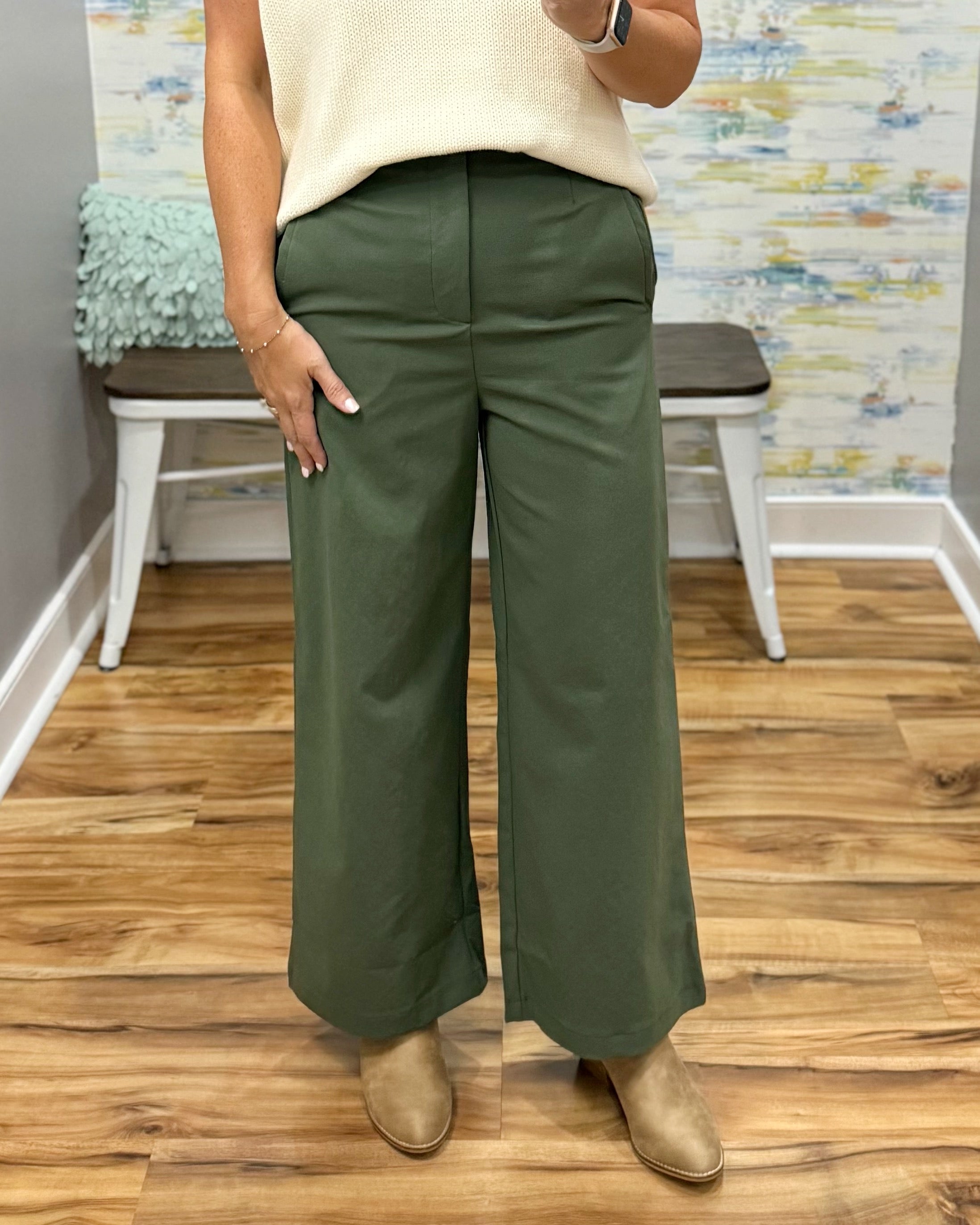 A person modeling a pair of wide-leg olive green pants with a cream knit top and tan ankle boots, standing on a wood floor in front of a bench and abstract wall art.