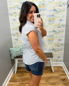 Woman taking a mirror selfie wearing a white sleeveless shirt and denim shorts in a room with a textured wall.