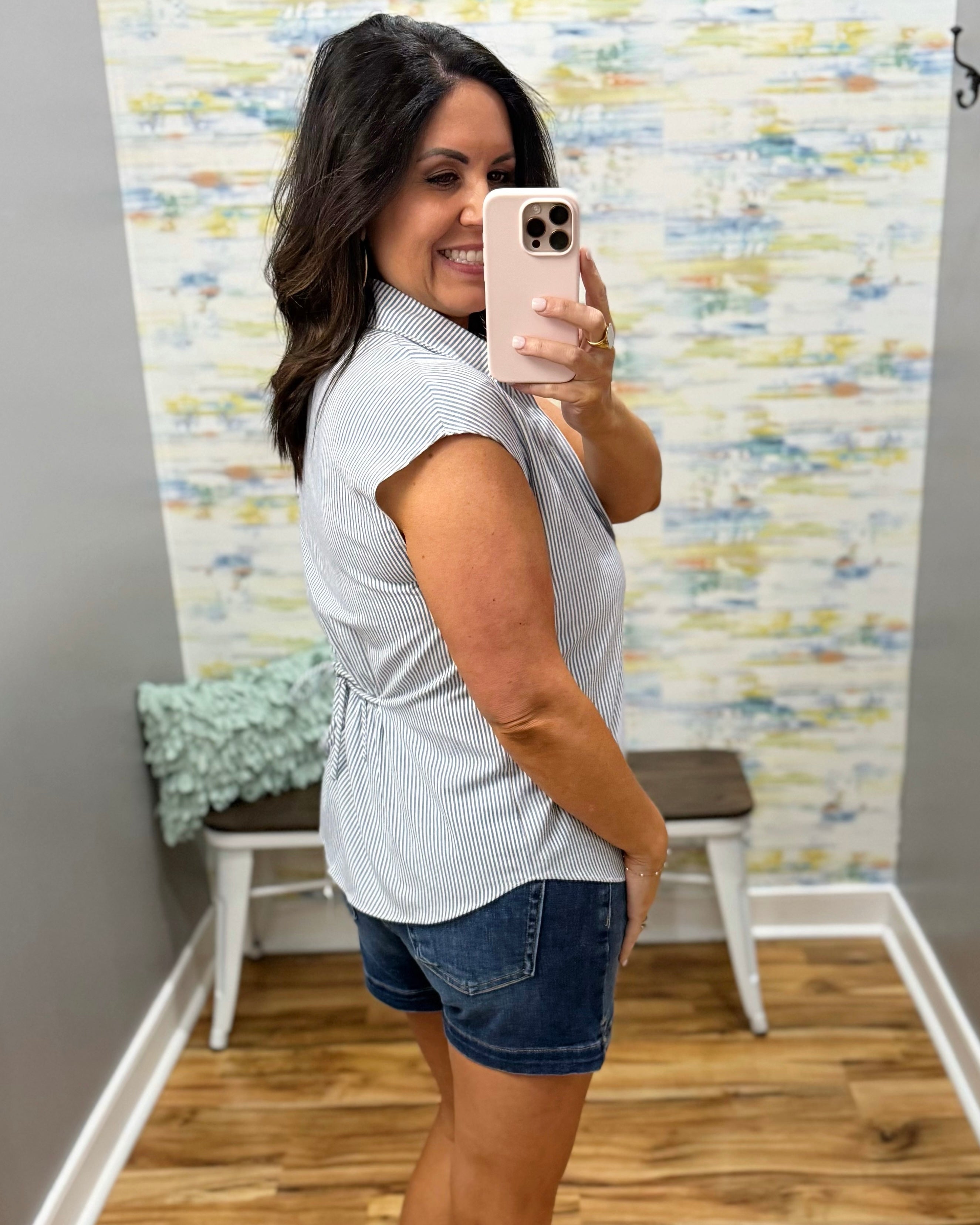 Woman taking a mirror selfie wearing a white sleeveless shirt and denim shorts in a room with a textured wall.