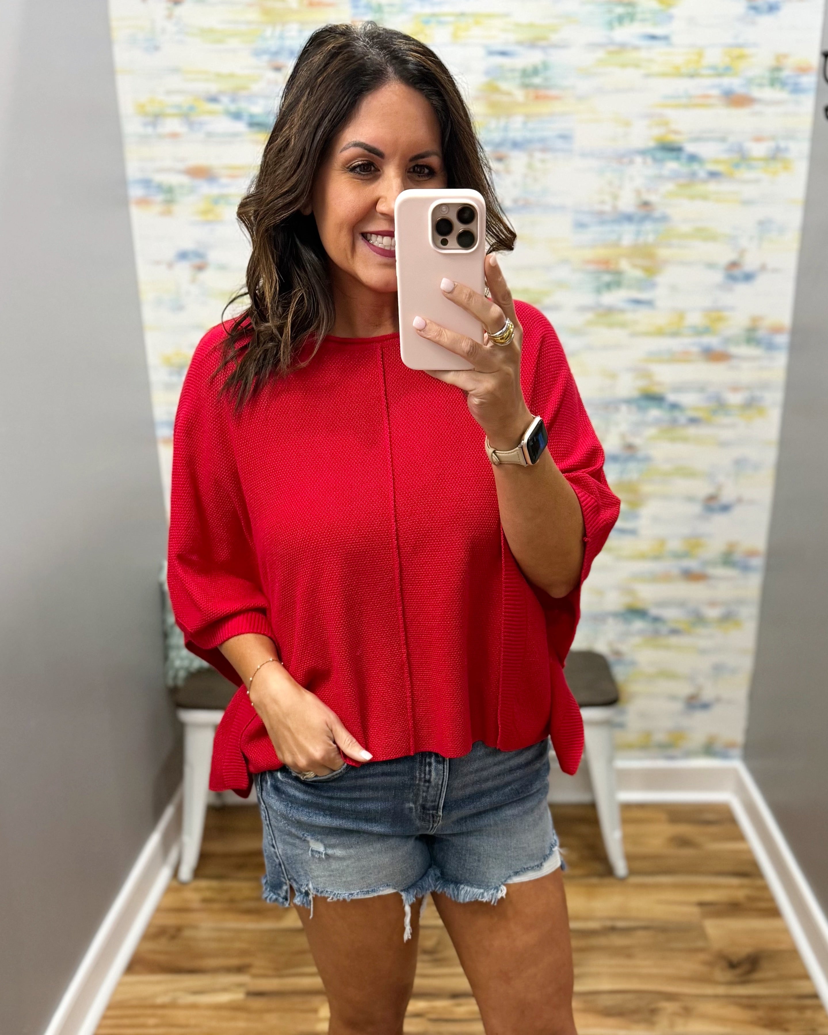 Woman taking a mirror selfie wearing a red top and denim shorts in a room with a colorful wall.