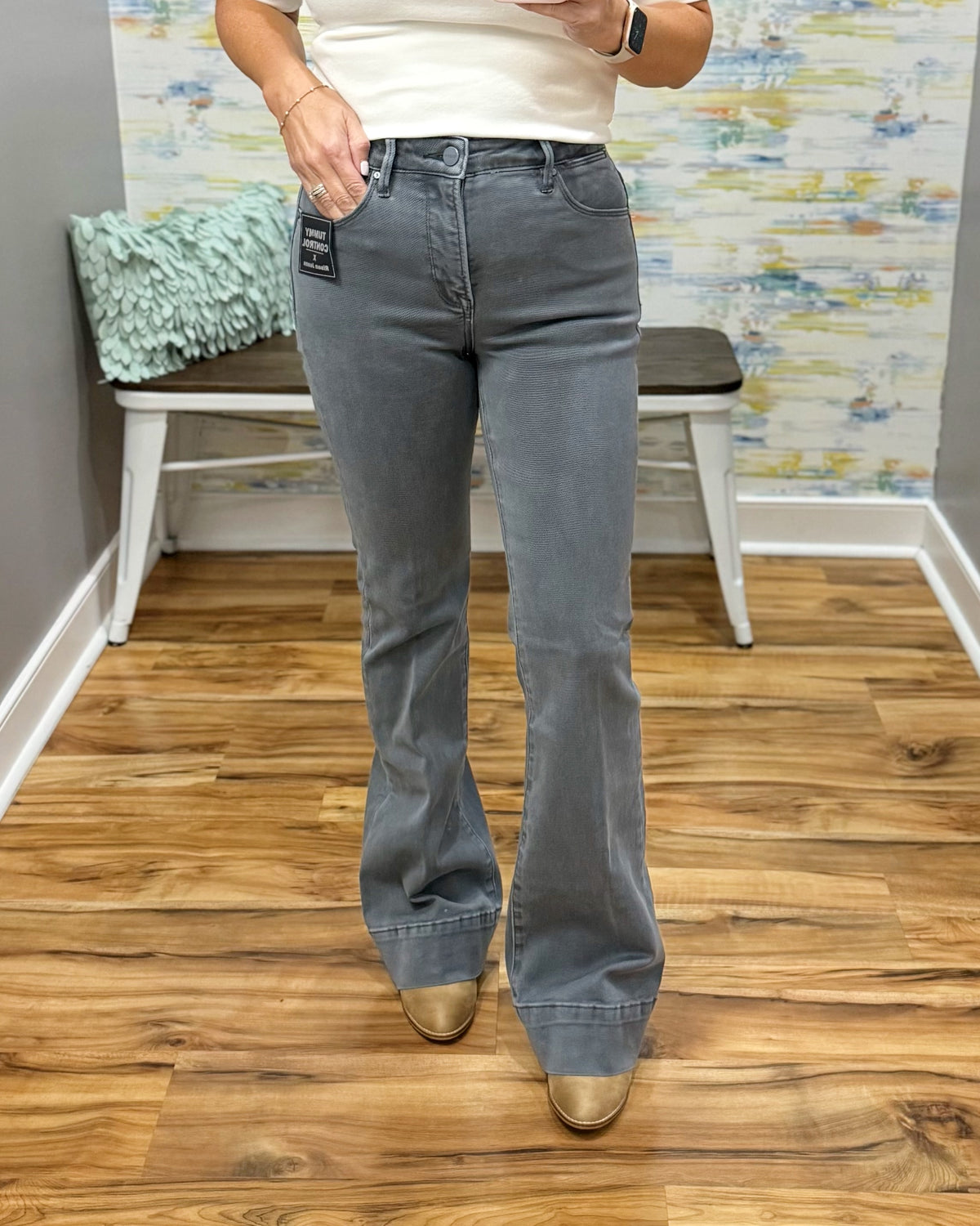 A person wearing medium-wash gray flared jeans and metallic gold boots, standing on a wood floor in front of a white bench and patterned wall.