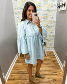 A woman takes a mirror selfie in a fitting room wearing a light blue chambray button-down tunic dress with flared bell sleeves and tan suede boots.