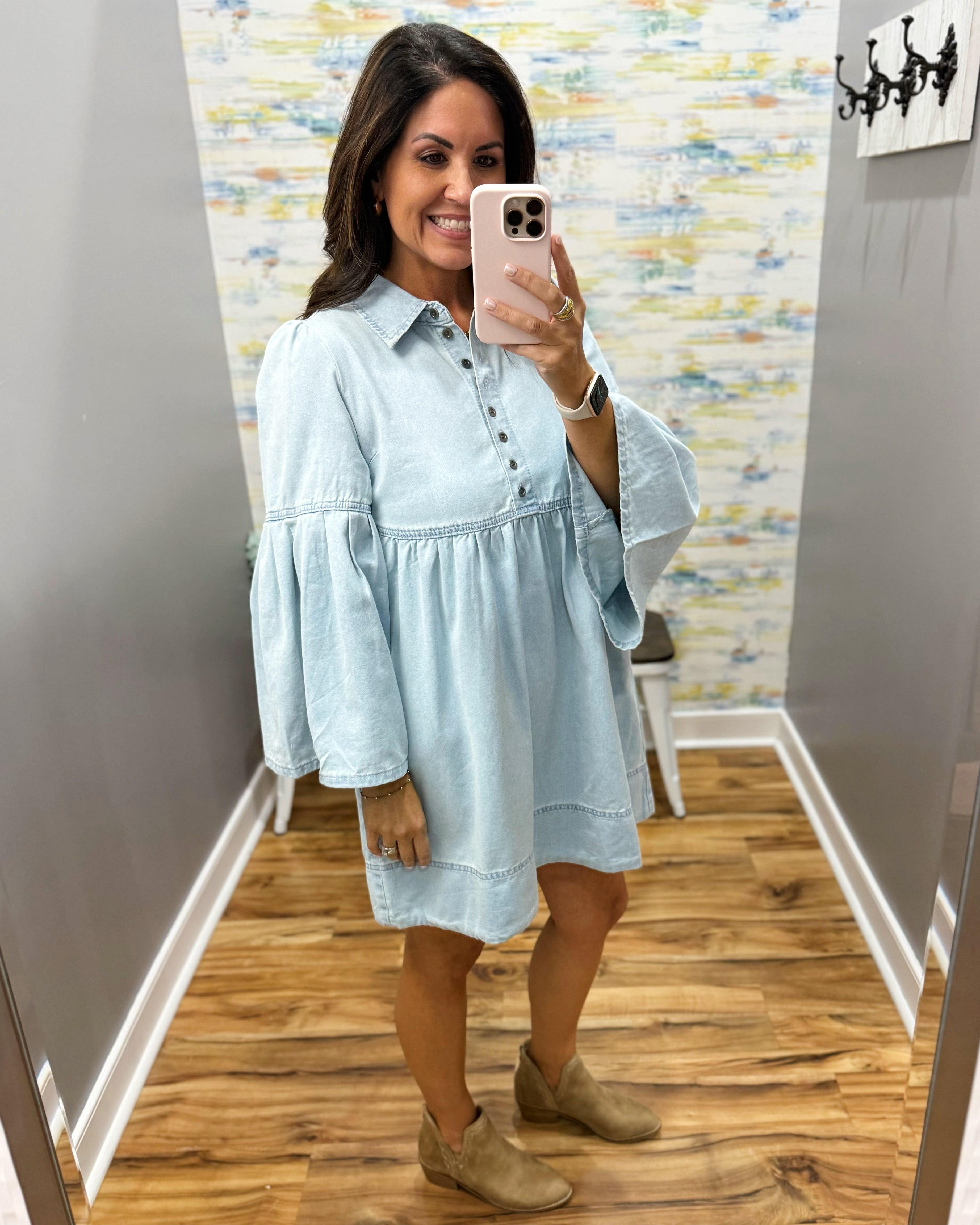 A woman takes a mirror selfie in a fitting room wearing a light blue chambray button-down tunic dress with flared bell sleeves and tan suede boots.