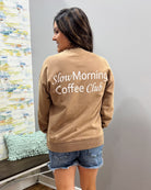 A woman shown from the back wearing a tan crewneck sweatshirt with 'Slow Morning Coffee Club' printed in white script across the back, paired with denim shorts in an indoor setting.