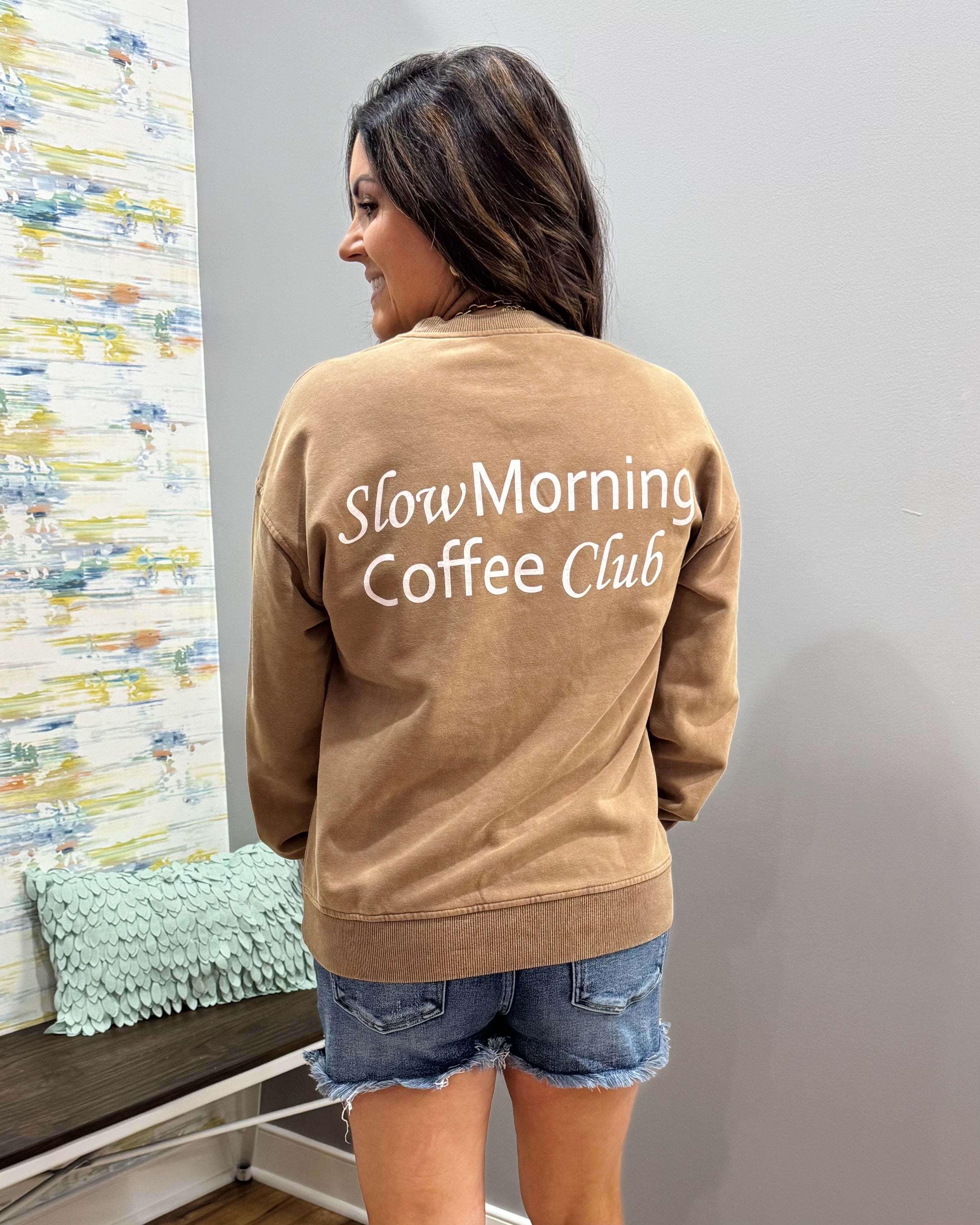 A woman shown from the back wearing a tan crewneck sweatshirt with 'Slow Morning Coffee Club' printed in white script across the back, paired with denim shorts in an indoor setting.
