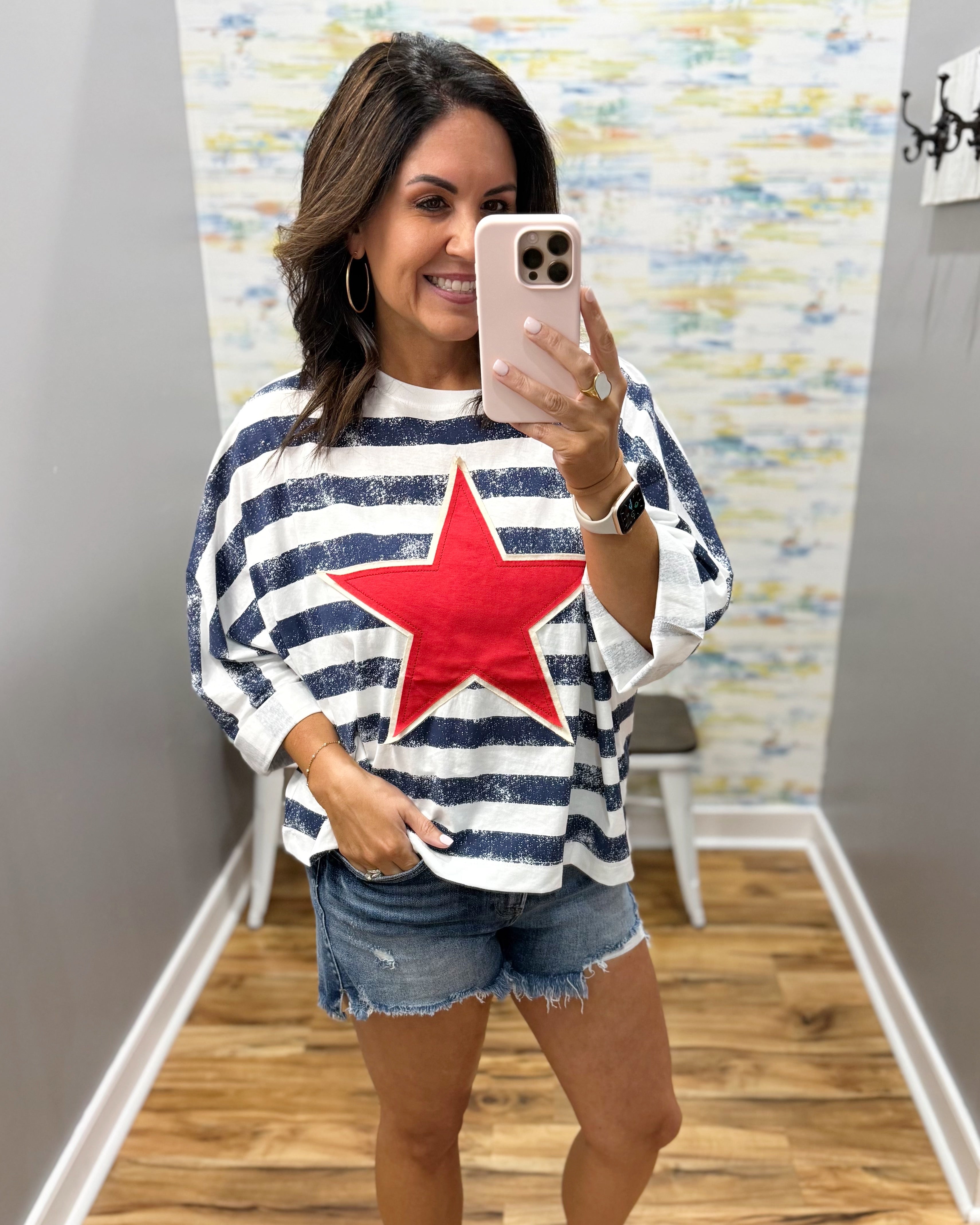 Woman taking a mirror selfie wearing a striped shirt with a red star and denim shorts.