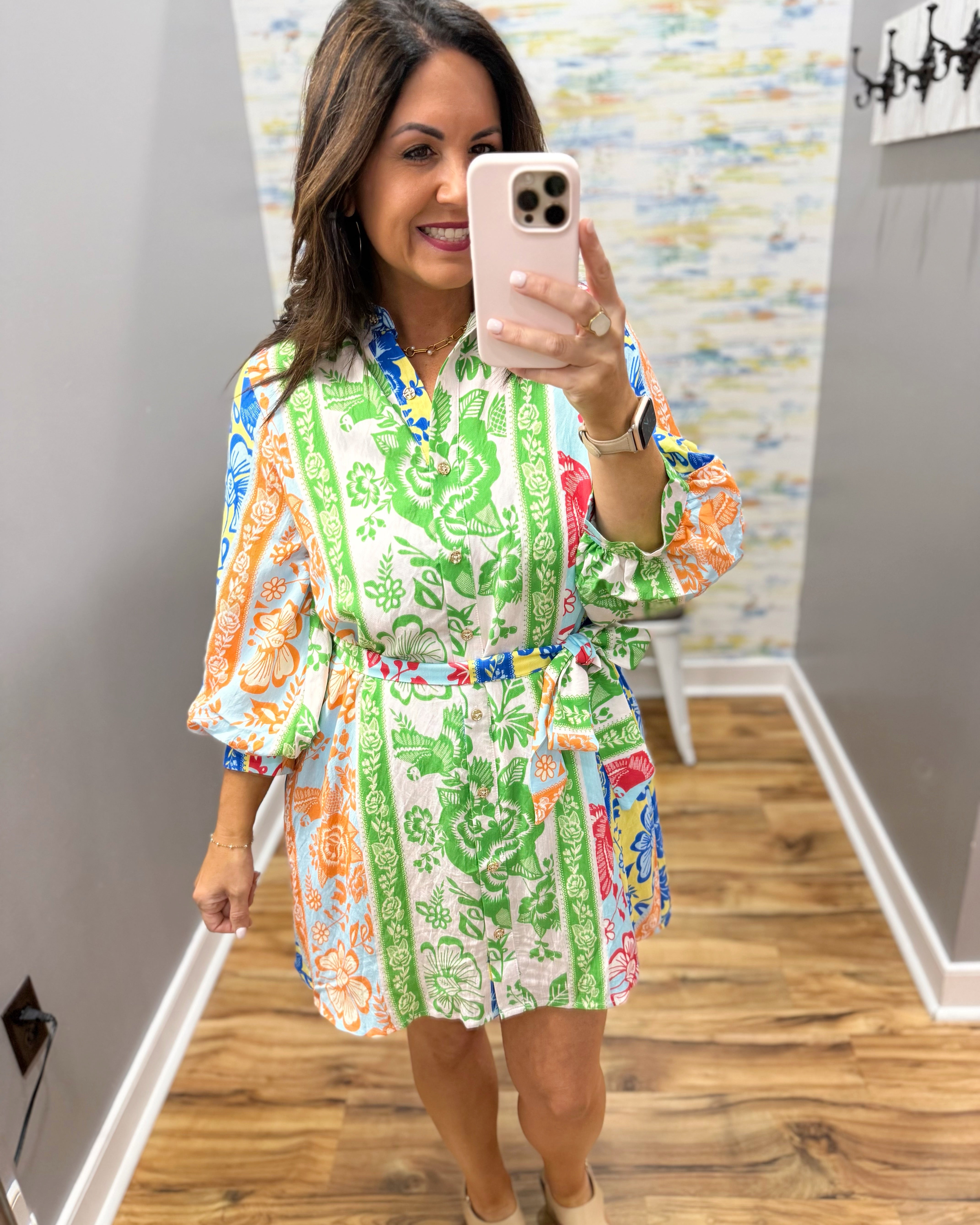 Woman taking a mirror selfie wearing a colorful floral dress in a room with a patterned wall.