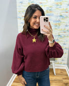 A woman takes a mirror selfie wearing a burgundy long-sleeve turtleneck sweater with balloon sleeves, paired with blue jeans and a gold star pendant necklace.