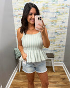 Woman taking a mirror selfie wearing a light green pleated top and denim shorts.