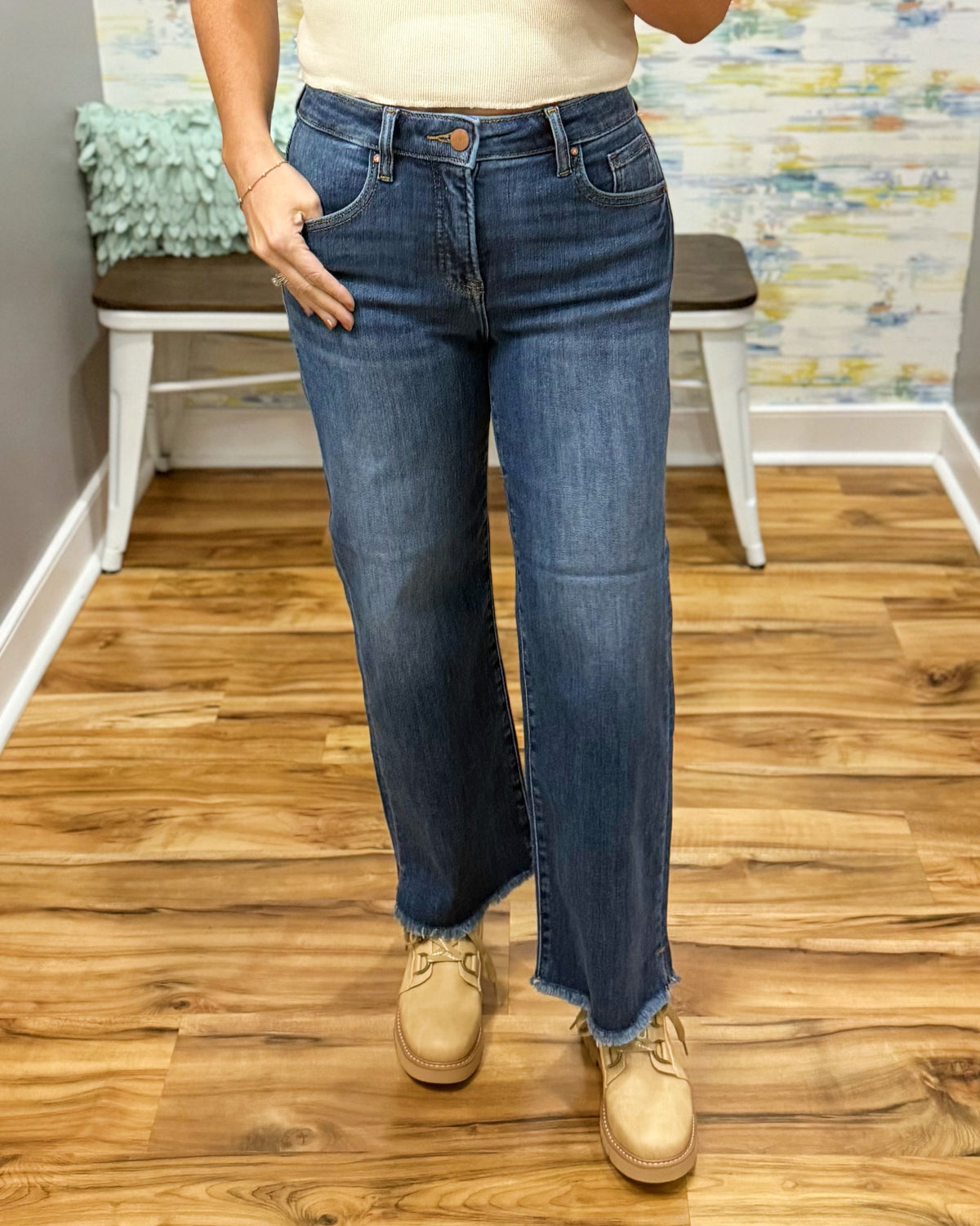 A person wearing blue Risen crop straight-leg jeans with a raw frayed hem, paired with beige boots and a cream top, standing on a wood floor in front of a white bench.