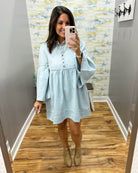 A woman takes a mirror selfie in a fitting room wearing a light blue long-sleeve button-up dress with a gathered waist and beige ankle boots.