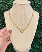 A gold chain necklace with a small interlocking ring clasp displayed on a beige fabric jewelry bust, with a hand holding the left side of the bust against a green leafy background.