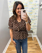A woman takes a mirror selfie wearing a brown short-sleeve blouse with white polka dots and blue jeans in a fitting room with wood floors and a patterned wall.
