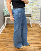 A person wearing mica wide-leg jeans, a black top, and tan pointed-toe heels, standing on a wood floor in an indoor room with a bench and abstract wall art in the background.