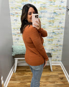 A woman takes a mirror selfie in a fitting room wearing a rust-orange ribbed knit sweater and light-wash blue jeans.