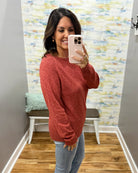 A woman takes a mirror selfie wearing a heathered rust-red knit sweater and light-wash blue jeans in a home setting with a patterned wallpaper background.