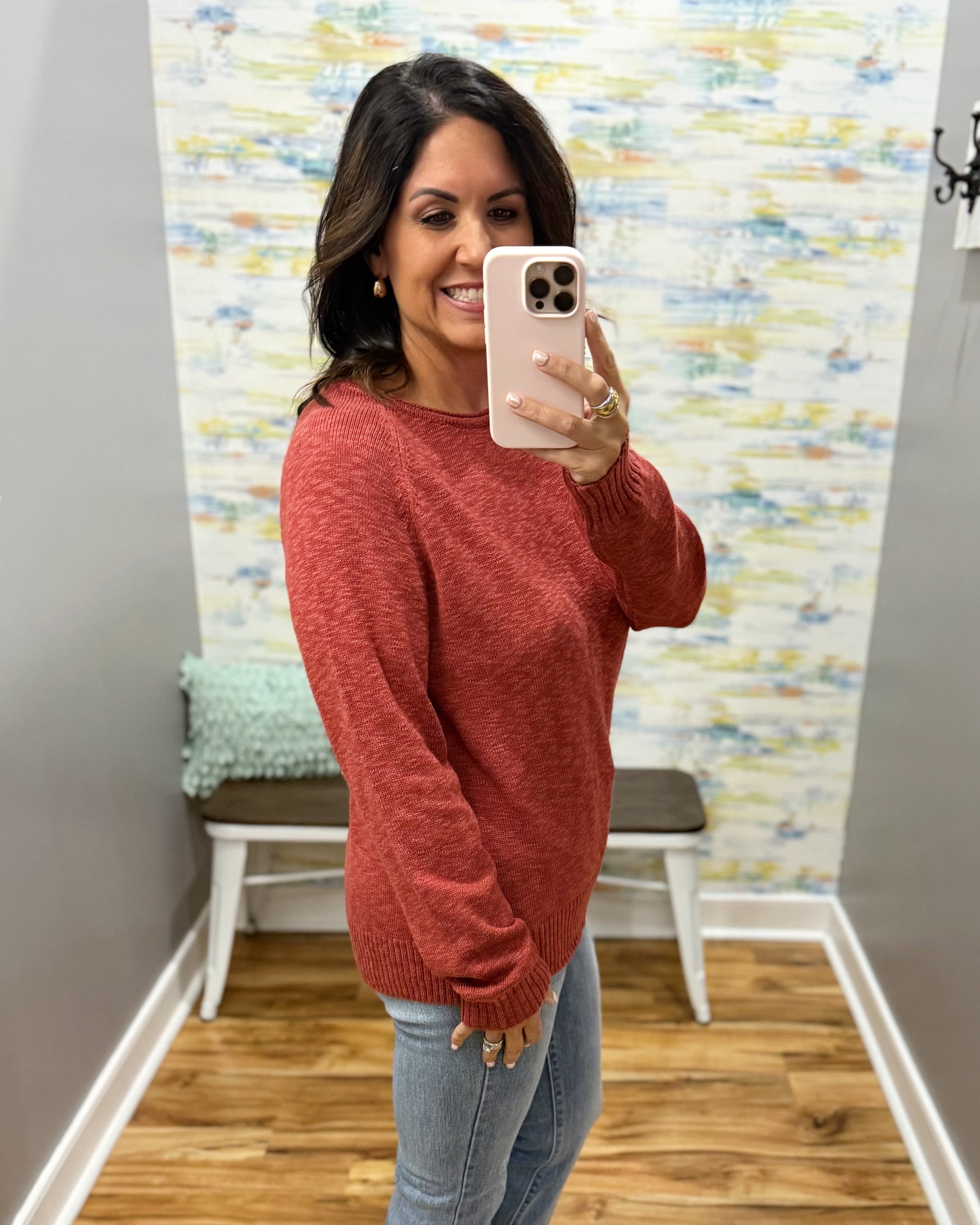 A woman takes a mirror selfie wearing a heathered rust-red knit sweater and light-wash blue jeans in a home setting with a patterned wallpaper background.