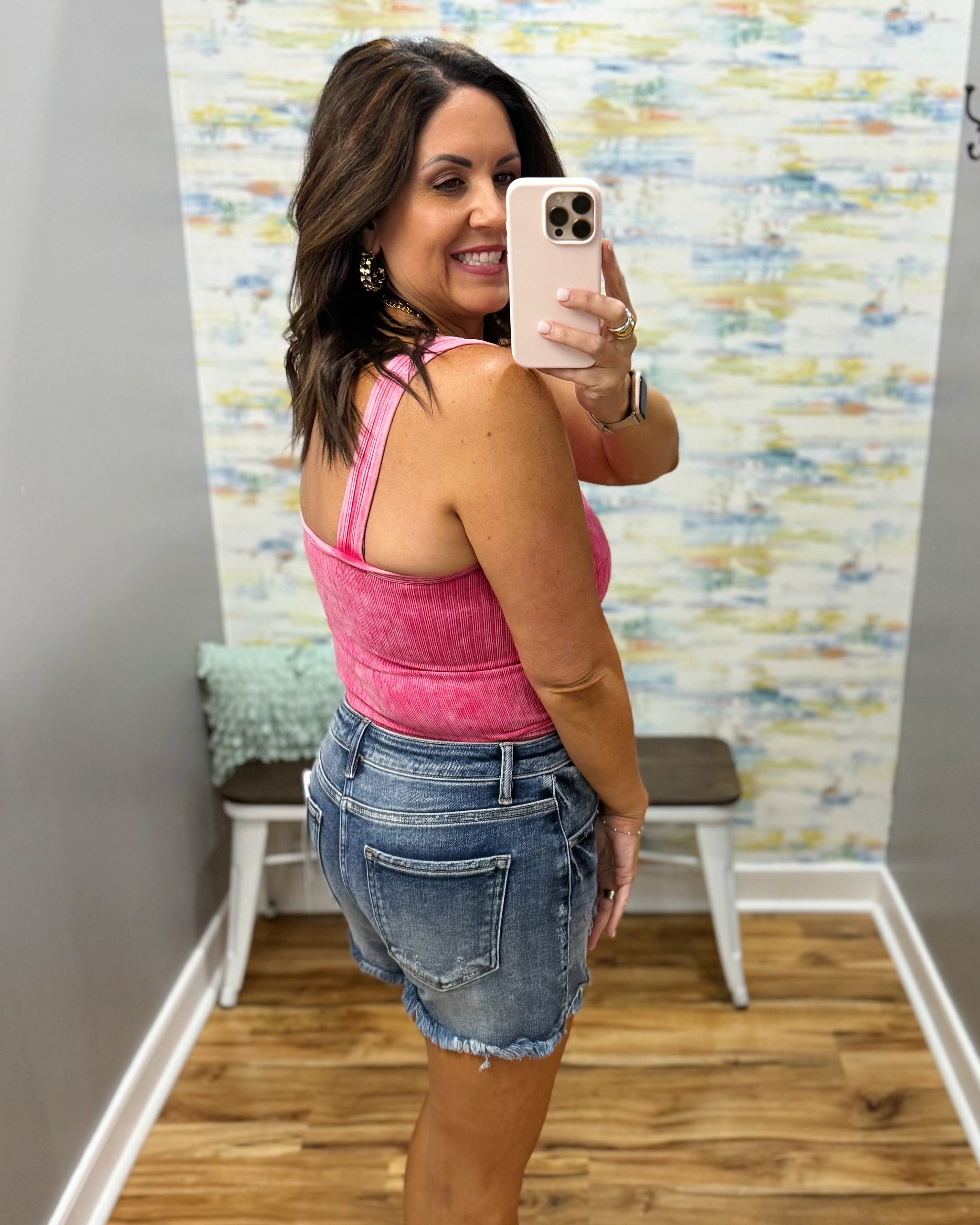 Woman taking a mirror selfie wearing a pink tank top and denim shorts in a room with colorful wall art.