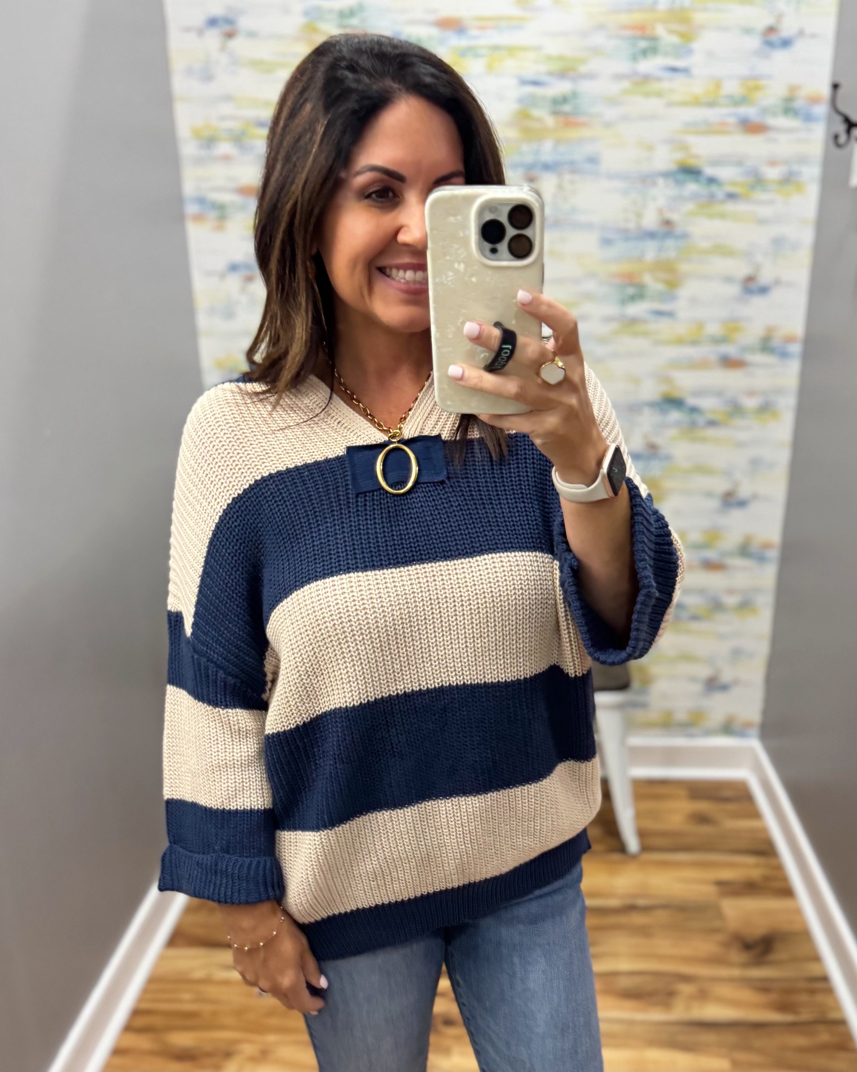 A woman takes a mirror selfie wearing a chunky knit sweater with horizontal navy blue and cream stripes, paired with blue jeans. The background shows a gray wall, a colorful abstract art print, and a light wood floor.