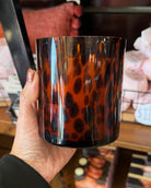 A hand holding a tall, cylindrical glass candle jar in sweet grace scent with a glossy amber-brown and black leopard print pattern, set against a blurred store background.