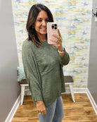 A woman takes a mirror selfie wearing a textured sage green knit sweater and light blue jeans in a home setting with a patterned wall and wood floor.