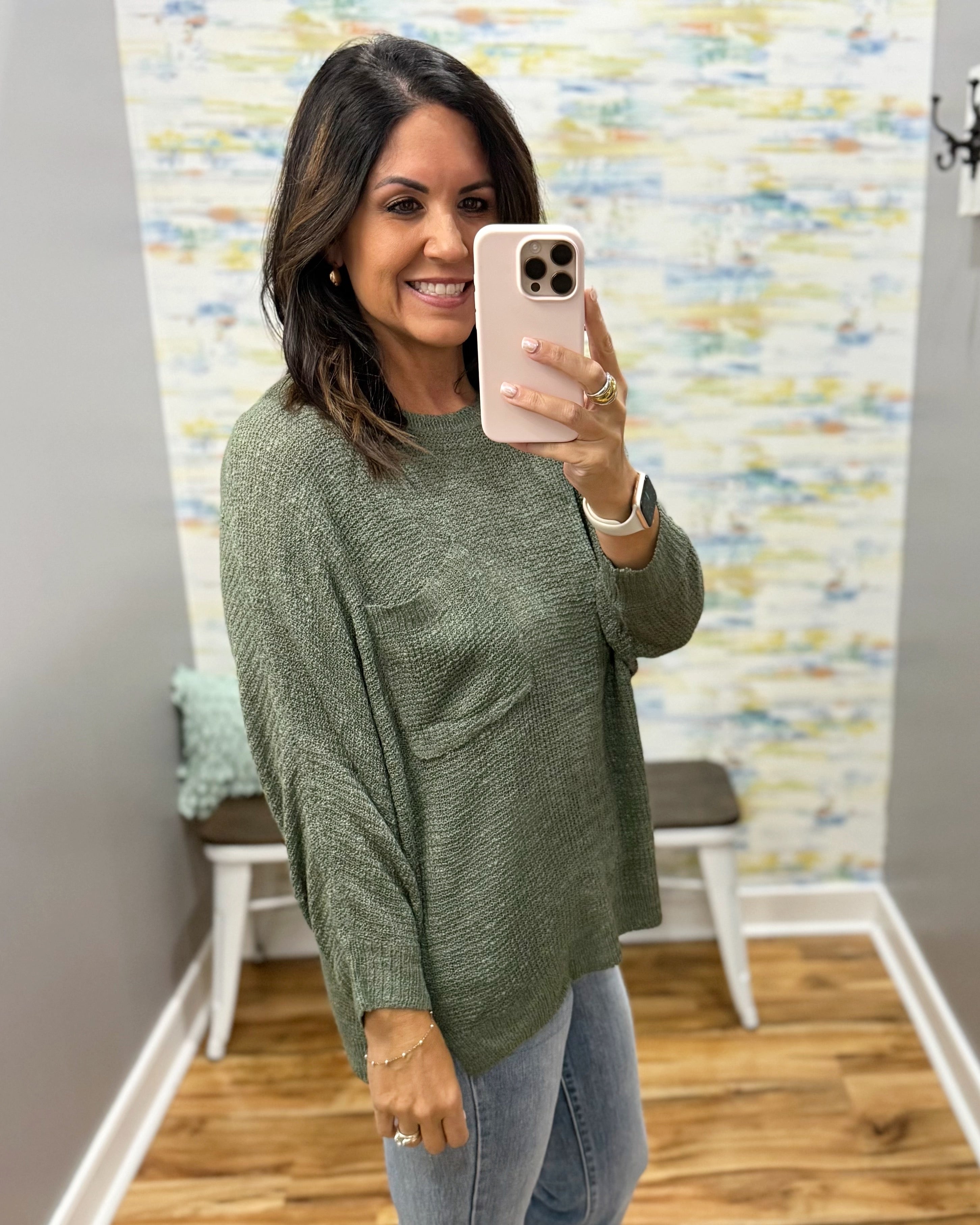 A woman takes a mirror selfie wearing a textured sage green knit sweater and light blue jeans in a home setting with a patterned wall and wood floor.