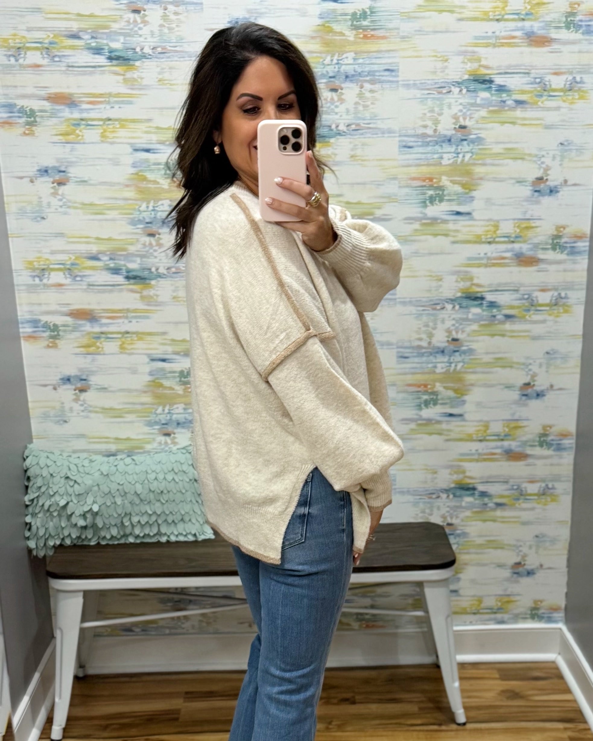 A woman takes a mirror selfie wearing a cream-colored, textured knit cardigan sweater and blue jeans in a fitting room with patterned wallpaper and a bench.