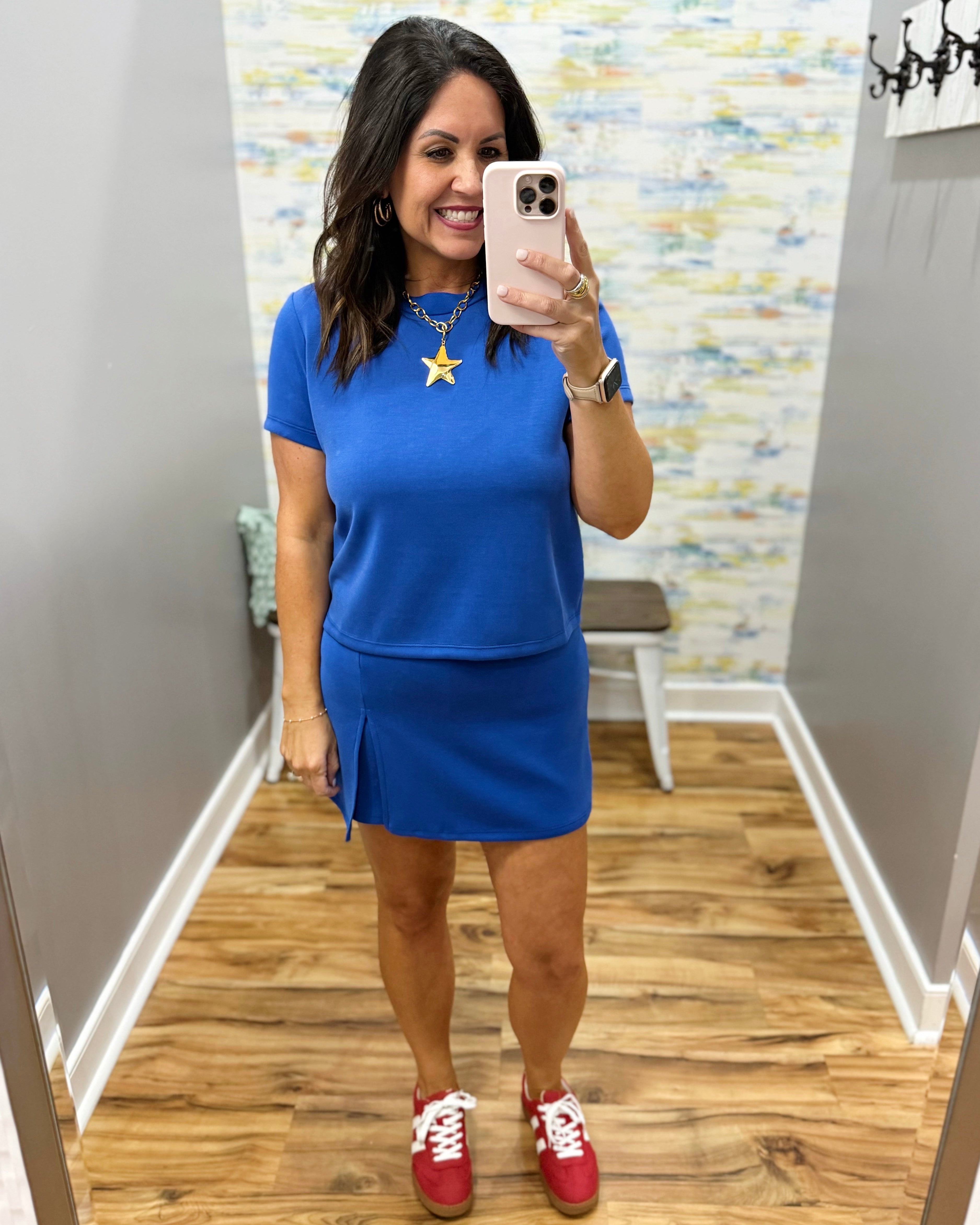Woman taking a mirror selfie wearing a blue outfit with red shoes in a room with patterned wallpaper.