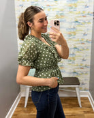 A woman takes a mirror selfie in a fitting room wearing an olive green short-sleeve blouse with white polka dots and dark blue jeans.