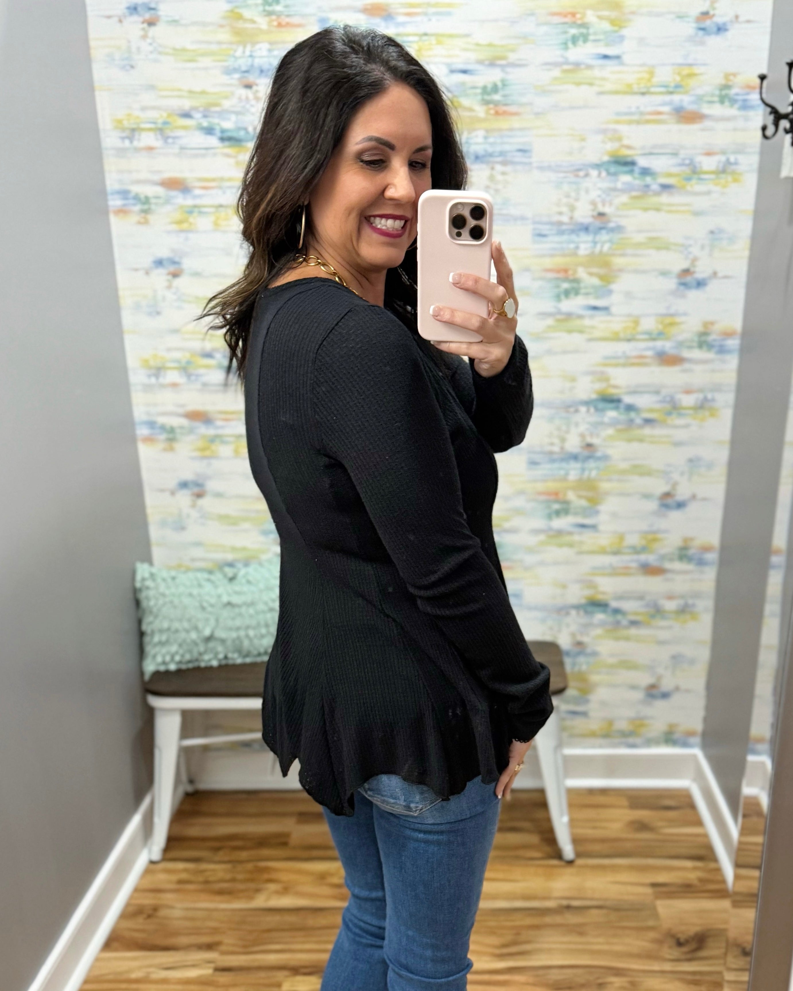 Woman taking a mirror selfie wearing a black peplum top and blue jeans in a room with patterned wallpaper.
