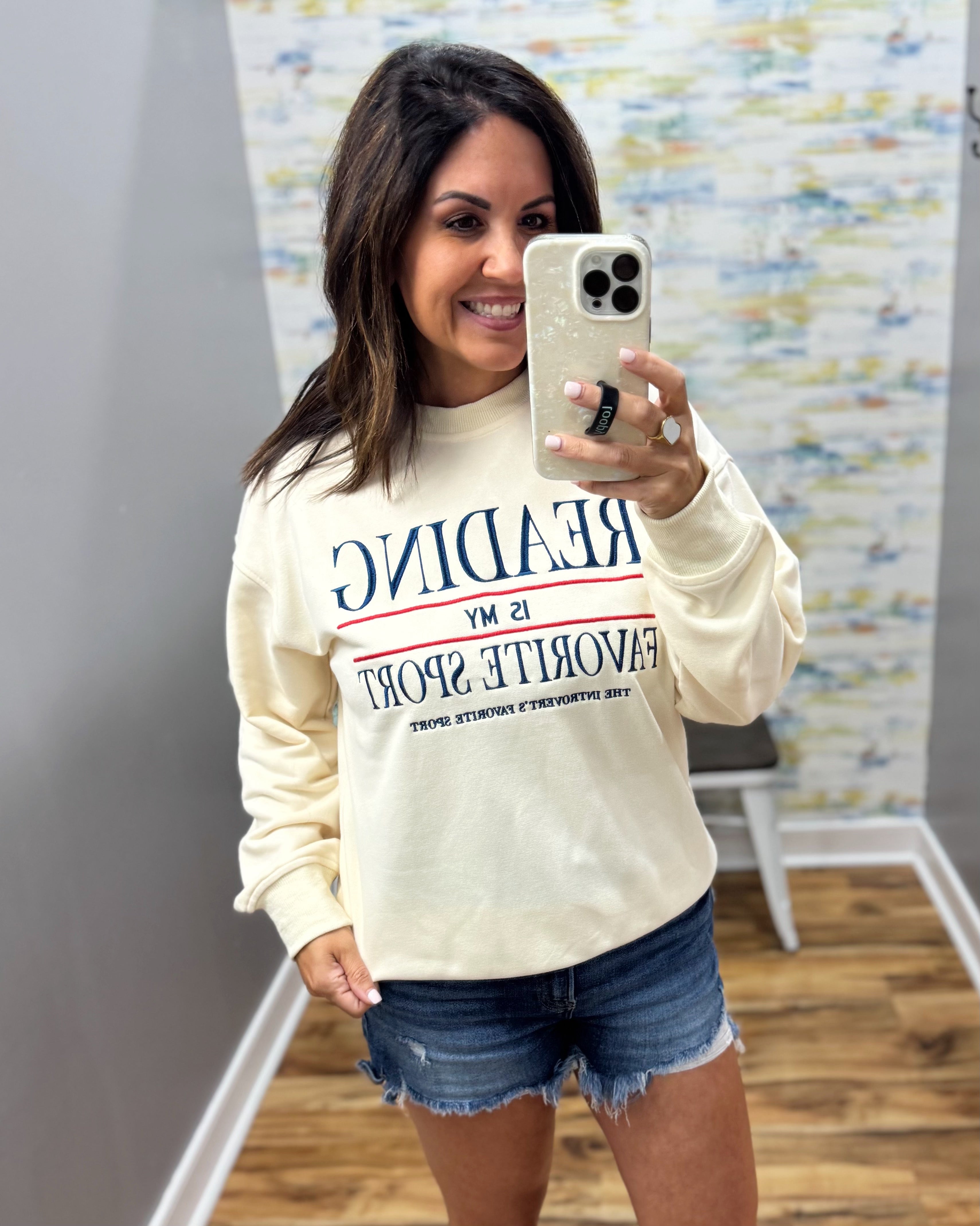 A woman taking a mirror selfie in a boutique wearing a cream-colored sweatshirt with blue text that reads 'READING IS MY FAVORITE SPORT' and denim shorts.