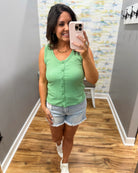 Woman taking a mirror selfie wearing a green tank top and denim shorts in a room with patterned wallpaper.