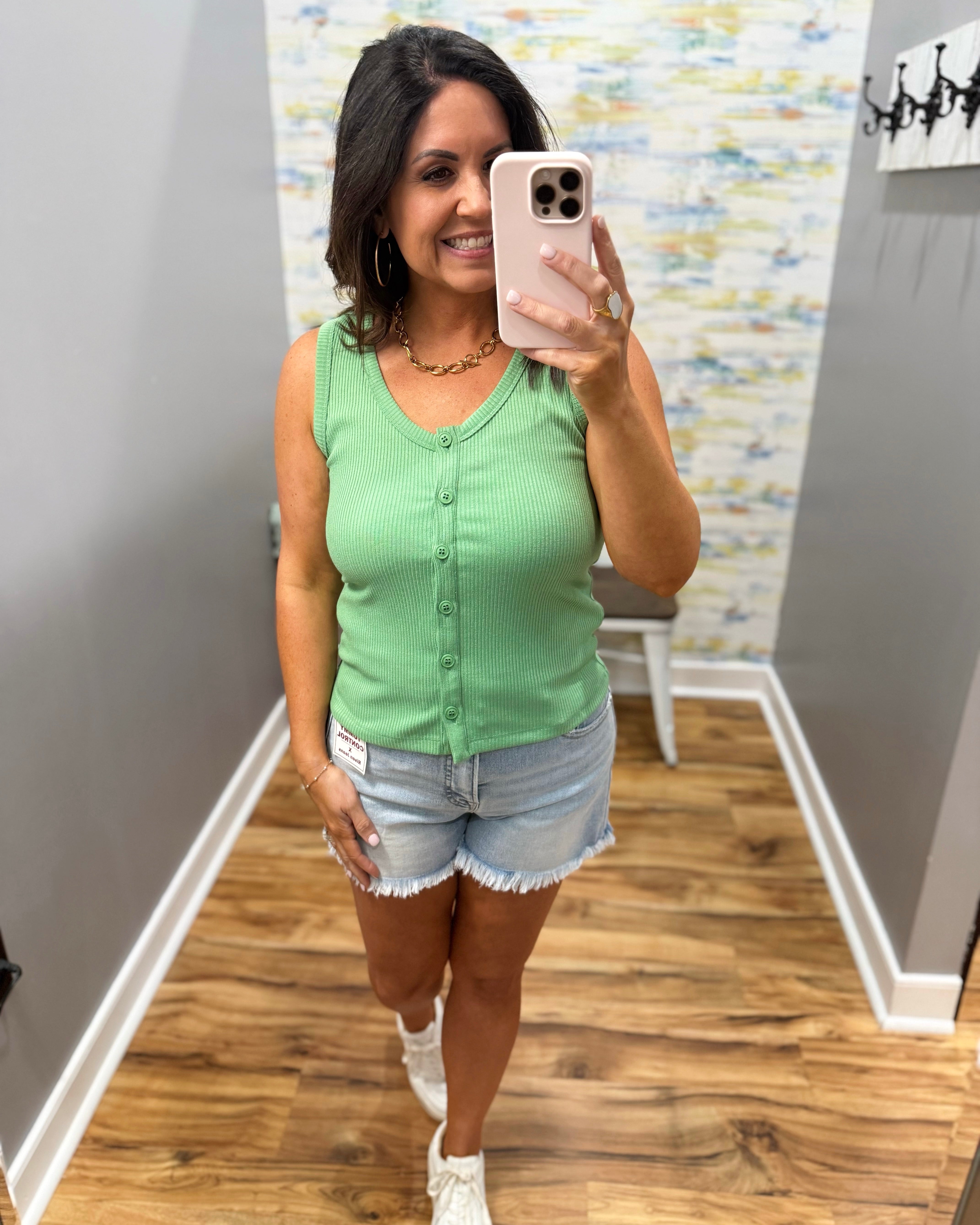 Woman taking a mirror selfie wearing a green tank top and denim shorts in a room with patterned wallpaper.